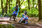 Two motocross riders in a forest trail, wearing helmets and blue gear, leaning into a turn on dirt bikes.