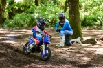 Two young motocross riders wearing helmets and protective gear ride dirt bikes on a wooded trail in sunlight.