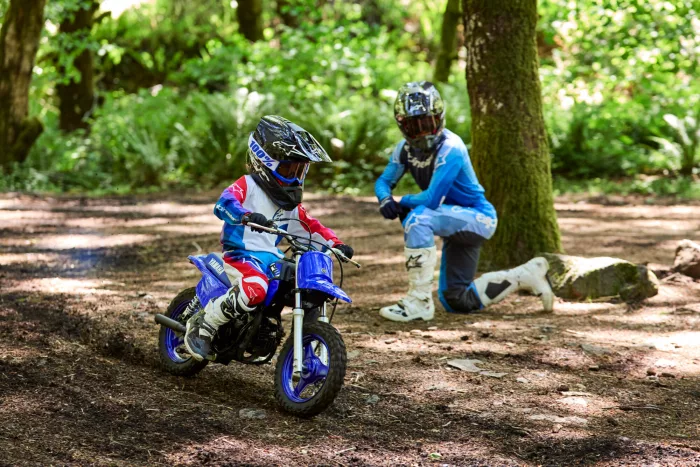 Two young motocross riders wearing helmets and protective gear ride dirt bikes on a wooded trail in sunlight.