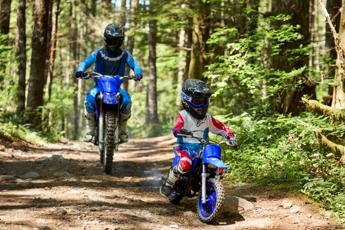 Two young dirt bike riders wearing helmets navigate a forest trail on their bikes.