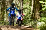 Child rider on a blue dirt bike leads two motocross riders through a sunlit forest trail with helmets on. (informative)