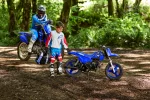 Two kids in full motocross gear with blue dirt bikes in a forest setting.