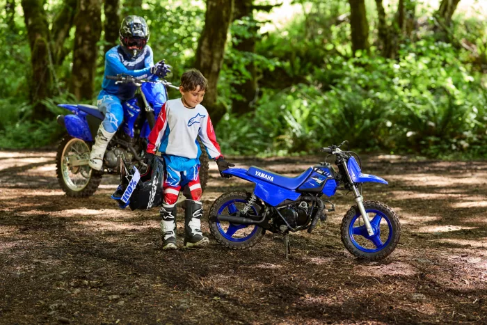 Two kids in full motocross gear with blue dirt bikes in a forest setting.