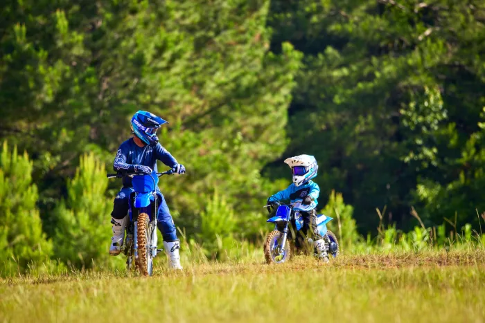 Two motocross riders in blue gear ride blue dirt bikes across a sunny grassy field with a green forest backdrop, helmets on.