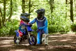Two young riders in motocross gear with helmets sit on a blue dirt bike in a forest clearing, a trainer kneels beside them.