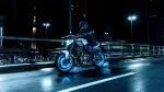 Motorcyclist in protective gear rides a white-and-blue bike on a wet city bridge at night with blue streetlights in the background.