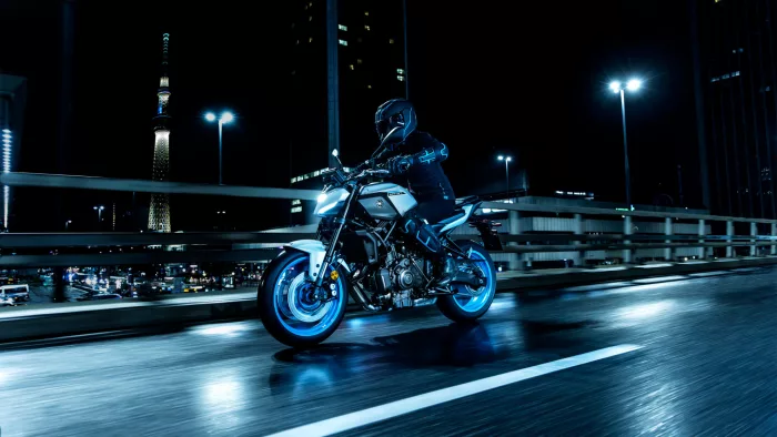 Motorcyclist in protective gear rides a white-and-blue bike on a wet city bridge at night with blue streetlights in the background.