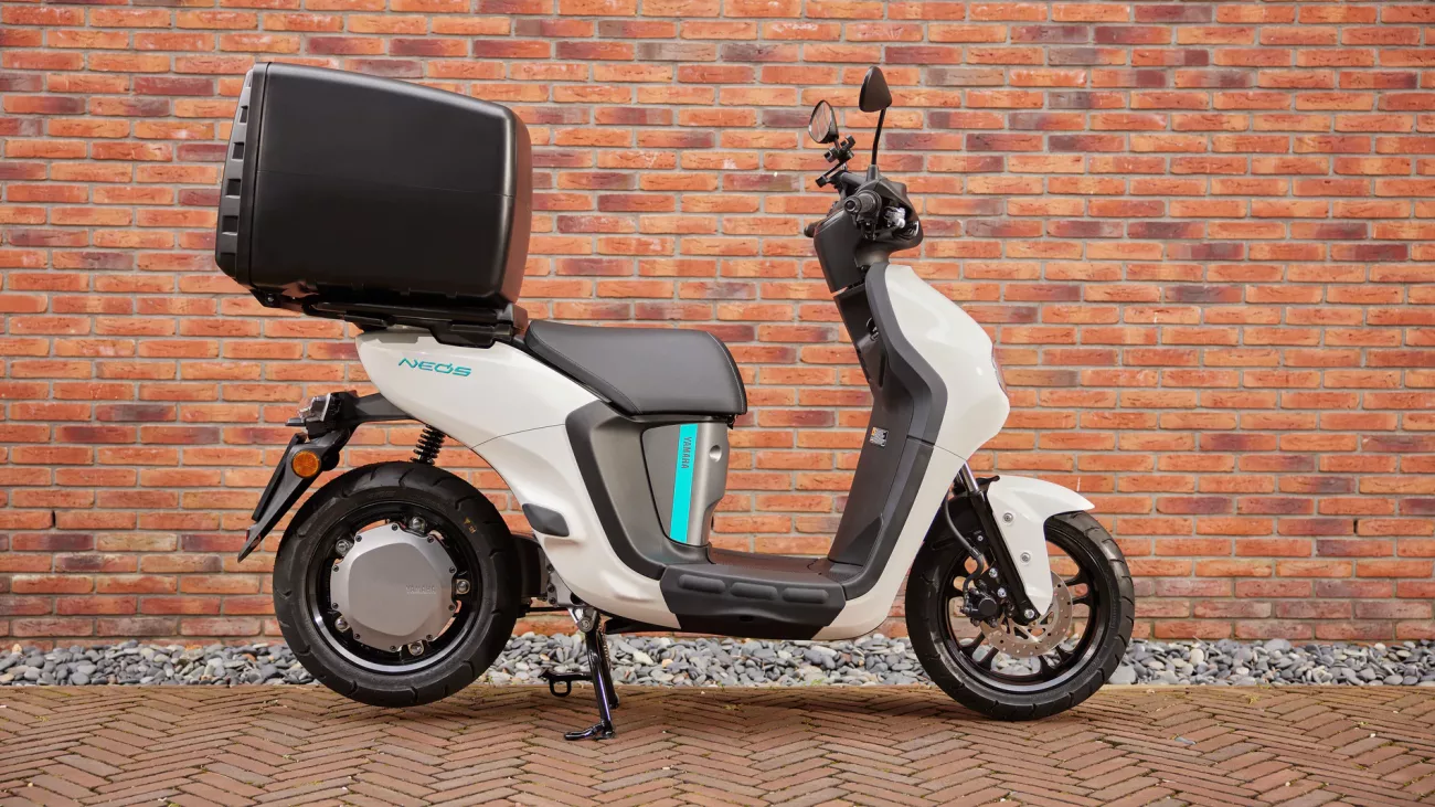 White Yamaha scooter with a large black top box parked on a brick wall backdrop.