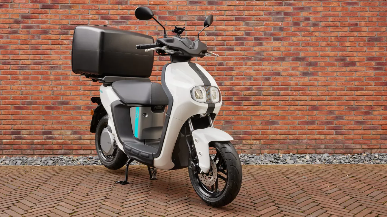 White and gray scooter with a large black top case parked on a brick-paved surface against a red brick wall, ready for commute or delivery use.