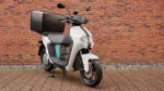White and gray scooter with a large black top case parked on a brick-paved surface against a red brick wall, ready for commute or delivery use.