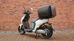 White Yamaha Neos scooter with a large black top case mounted on the rear, parked on a brick-paved surface with a brick wall backdrop.