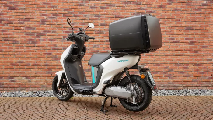 White Yamaha Neos scooter with a large black top case mounted on the rear, parked on a brick-paved surface with a brick wall backdrop.