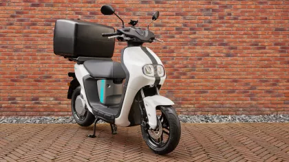 White and gray scooter with a large black top case parked on a brick-paved surface against a red brick wall, ready for commute or delivery use.