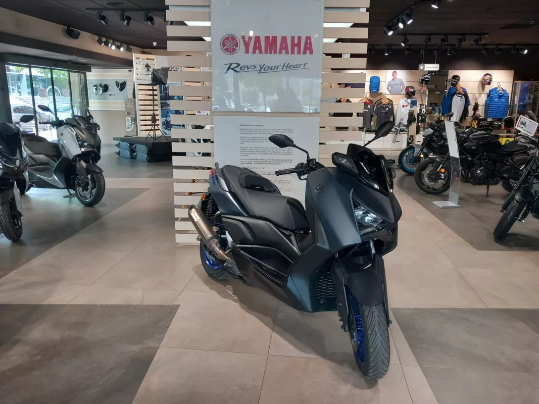 Matte dark Yamaha sport motorcycle in the showroom foreground with other bikes and apparel displays in the background, Yamaha sign reading 'Revs Your Heart' visible.