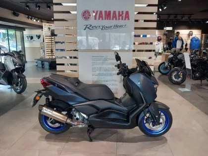 Inside a Yamaha showroom, a dark blue Yamaha XMAX scooter in the foreground with other bikes, helmets, and gear in the background.
