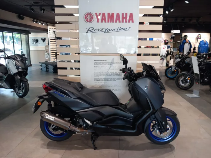 Inside Yamaha dealership showroom with a large Yamaha sign and 'Rev Your Heart' banner; foreground features a dark-blue sport scooter on the floor.
