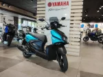 Blue Yamaha scooter on showroom floor with other bikes and a large Yamaha sign in the background, indoors showroom atmosphere.