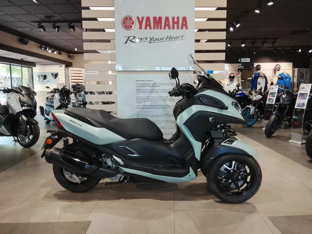 Teal Yamaha Tricity scooter on display in a motorcycle showroom, with other bikes in the background.