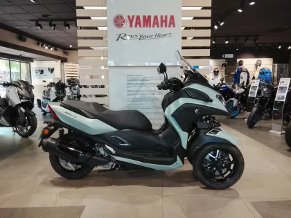 Teal Yamaha Tricity scooter on display in a motorcycle showroom, with other bikes in the background.