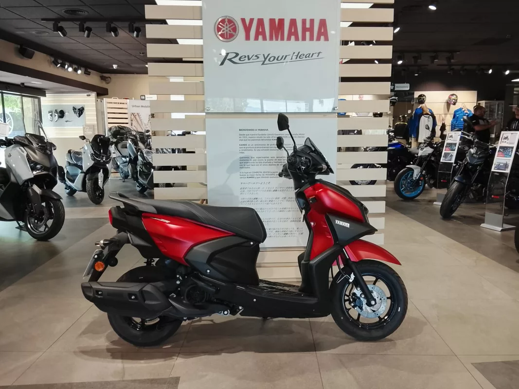 Red Yamaha scooter in the foreground of a brightly lit motorcycle showroom with other bikes on display and a central display pillar behind it.