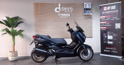 Blue Yamaha scooter on a showroom floor, with a wooden slat wall and a promotional banner nearby.