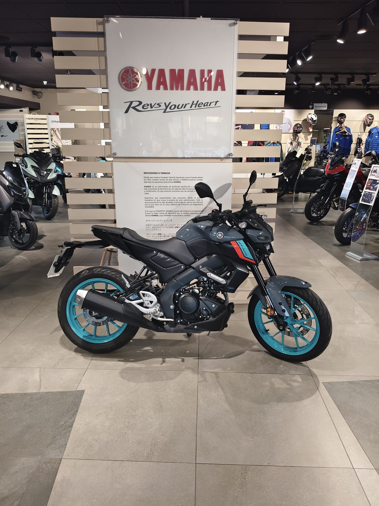 Black Yamaha motorcycle with turquoise rims on a showroom floor, large Yamaha logo and gear displays in the background.